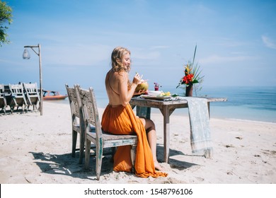 Back View Of Pleased Blond Lady In Orange Dress Dreaming While Sitting Alone At Rustic Served Table On Old Chair And Having Fresh Coconut Drink On Coast At Bali Resort Against Tranquil Ocean Waves