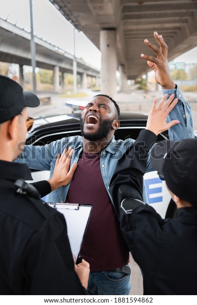back\
view of police officers calming despaired african american man near\
patrol car on blurred background on urban\
street