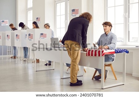 Back view portrait american man with long hair registering at polling station with American flags on election day. Young USA citizen voter standing at vote center and getting ballot paper.