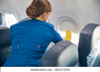 Back View Portrait Of Beautiful Charming Female Wearing Fight Attendant Blue Uniform Closing Illuminator Near The Cozy Passenger Chairs