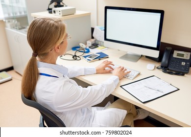 Back View Portrait Of Blonde Female Doctor Sitting At Desk In Office Working With Computer, Typing And Looking At Blank White Screen, Copy Space