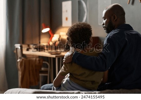 Back view portrait of caring single father embracing son sitting on bed together and talking copy space