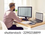 Back view portrait of a concentrated young business man accountant sitting at the desk and working on a laptop with tables and charts at office or at home and looking to the monitor screen.