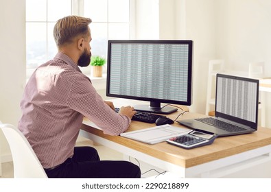Back view portrait of a concentrated young business man accountant sitting at the desk and working on a laptop with tables and charts at office or at home and looking to the monitor screen.