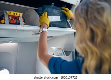 Back View Portrait Of Friendly Air Hostess In Yellow Gloves Putting Handbag In The Cabin Compartment