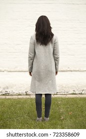 Back View. Portrait Of A Full Length Brunette Woman In A Gray Woolen Coat In A Stone Wall Of White Brick