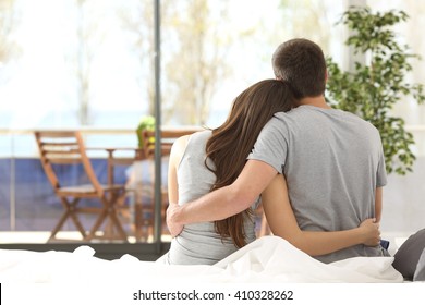 Back View Portrait Of A Happy Couple Sitting On The Bed Looking The Balcony Outdoors Through A Window Of The Bedroom Of A House