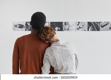Back view portrait of mixed-race couple embracing while looking at paintings at modern art gallery exhibition, copy space