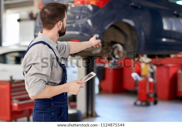 Back view portrait of modern bearded mechanic\
holding digital tablet pointing at car while working in car service\
and repair center, copy\
space