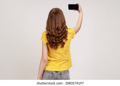 Back View Portrait Of Slim Female In Yellow Casual T-shirt Talking Video Call, Taking Selfie On Mobile Phone, Holding Smartphone Up With Blank Display. Indoor Studio Shot Isolated On Gray Background.