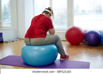 Back View Portrait Of Sweaty Overweight Woman Working Out In Fitness Studio: Sitting On Big Fitness Hanging Her Head Looking Upset And Exhausted