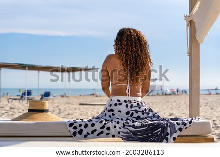 Back view portrait of unrecognizable afro latin female with beautiful stylish pareo sitting on a bench on beach landscape.