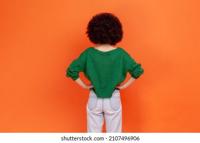 Back View Portrait Of Woman With Afro Hairstyle Wearing Green Casual Style Sweater Standing Posing Backwards, Keeping Hands On Hips. Indoor Studio Shot Isolated On Orange Background.