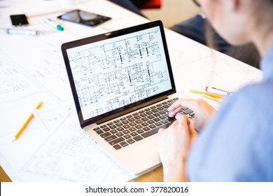 Back View Portrait Of A Woman Engineer Working On Laptop Computer With Blueprints On Screen In Office