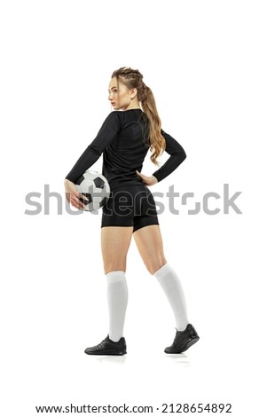 Back view. Portrait of young beautiful girl, professional female soccer player holding football ball isolated on white background. Concept of sport, fitness. Young spotive girl in black football kit