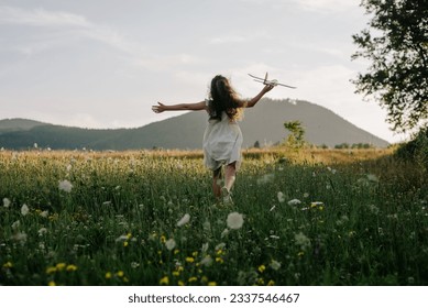 Back view of preteen girl in dress playing with violet toy airplane during beautiful warm sunset. Childhood dreams. Family, nature, freedom and airplane concept. Playful cute little child pilot
