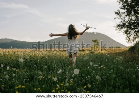 Back view of preteen girl in dress playing with violet toy airplane during beautiful warm sunset. Childhood dreams. Family, nature, freedom and airplane concept. Playful cute little child pilot