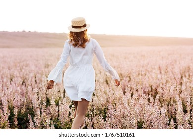 Back View Of A Pretty Young Woman In White Dress Holding Straw Hat Running Walking At The Sage Flower Field. Beautiful Girl Enjoying A Field Of Flowers, Relaxing Outdoors, Having Fun, Harmony Concept.