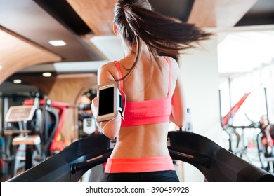 Back View Of Pretty Young Woman Athlete With Balnk Screen Smartphone Running On Treadmill In Gym