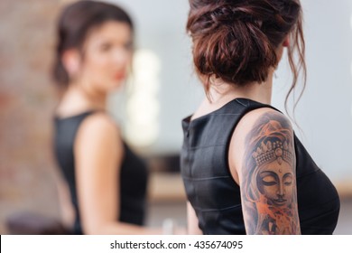 Back View Of Pretty Young Woman In Black Dress With Tattoo On Her Hand Standing In Front Of The Mirror