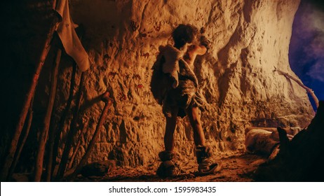 Back View Of Primitive Prehistoric Neanderthal Wearing Animal Skin Draws Animals And Abstracts On The Walls At Night. Creating First Cave Art With Petroglyphs, Rock Paintings Illuminated By Fire