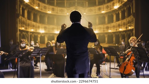 Back View of Professional Conductor Directing Symphony Orchestra with Performers Wearing Medical Masks, Playing Violins, Cello and Trumpet on Classic Theatre with Curtain Stage During Music Concert