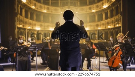 Back View of Professional Conductor Directing Symphony Orchestra with Performers Wearing Medical Masks, Playing Violins, Cello and Trumpet on Classic Theatre with Curtain Stage During Music Concert Foto stock © 