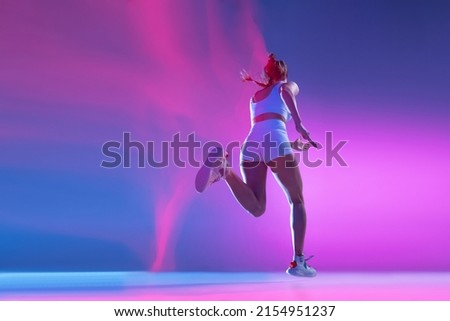 Back view. Professional female athlete running away isolated on blue studio background in mixed pink neon light. Sportive girl in white sportswear practicing in run. Healthy lifestyle concept.