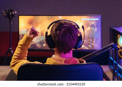 Back view of professional gamer sitting in gaming chair and celebrating his victory in online esport tournament, makes winner hand gesture, wearing headset, looking on monitor. Neon light. Cyber sport