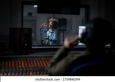 Back View Of Professional Male Sound Producer With Headphones Is Recording A New Song With Young African Female Singer In A Music Studio.