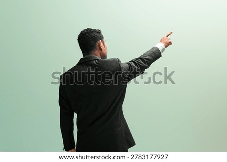 Back view of a professional man in a black suit pointing finger up and forward against a clean green background with blank space.