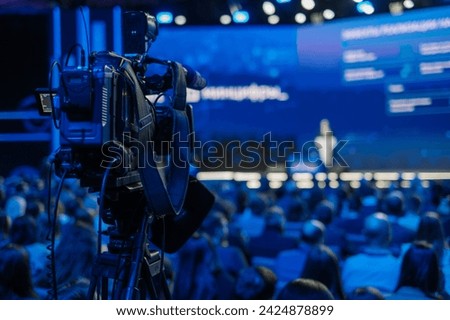 Back view of a professional video camera recording a speaker at a crowded business conference with audience.