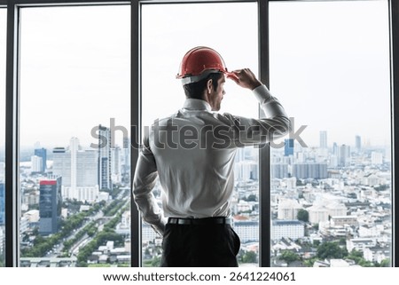 Back view project manager man with safety helmet standing in high rise office looking out of window in downtown cityscape