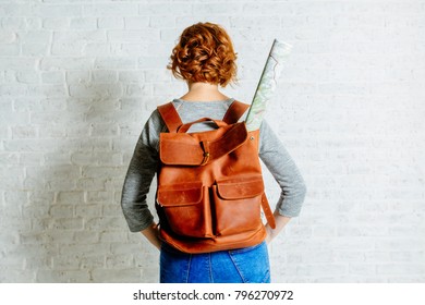 Back view of red hair woman in gray shirt and blue jeans and red brown leather backpack with a rolled roadmap over white brick wall with shadow backround. Travel concept.