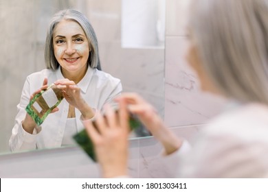 Back View, Reflection In The Mirror Of Attractive Senior Gray Haired Woman Applying Moisturizing Antiwrinkle Facial Mask On Her Face While Standing In Front Of The Mirror In Her Bathroom At Home