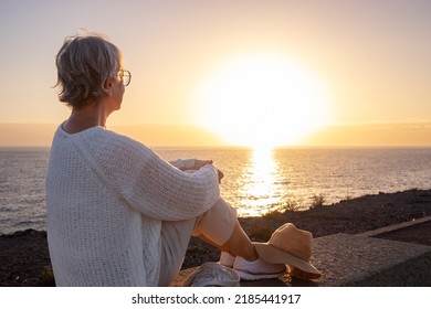 Back view of relaxed and romantic senior woman or pensioner sitting admiring the sea at sunset light looking at the horizon - old female  outdoors enjoying vacations - Powered by Shutterstock