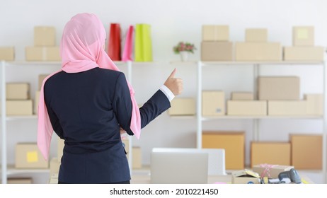 Back view religious asian muslim woman in blue suit and pink shaft on head standing with confidence showing thum up. Business woman stand with package SME box delivery background. Work at home concept
