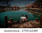 Back view of romantic pair embracing on pier watching boats and tropical resort buildings reflected in turquoise lagoon water under cloudy sky.