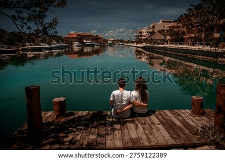 Back view of romantic pair embracing on pier watching boats and tropical resort buildings reflected in turquoise lagoon water under cloudy sky.