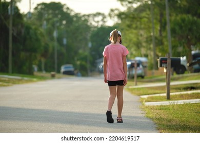 Back View Of Sad Young Child Girl Walking Alone Along The Green Street On Sunny Summer Day