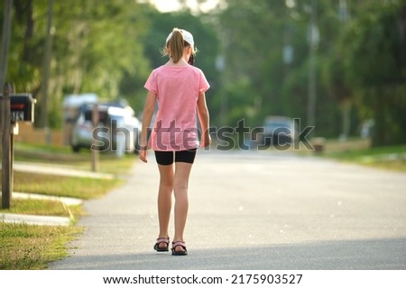 Back view of sad young child girl walking alone along the green street on sunny summer day