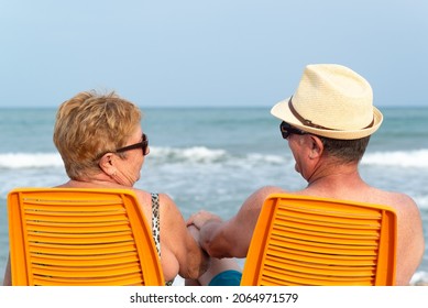 Back view of a senior couple in love looking at each other and holding hands while they are sitting at the beach. Relaxed summer vacation.