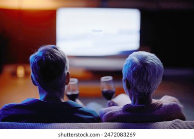 Back view of senior couple watching TV together sitting on sofa at home with hands holding wine glasses copy space - Powered by Shutterstock