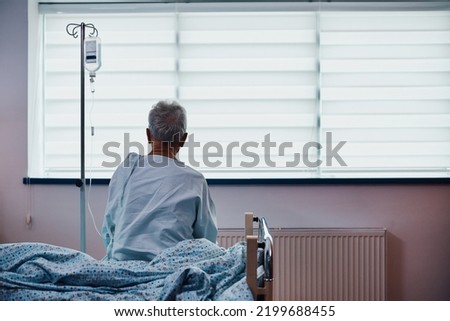 Back view of senior male patient with IV drip sitting on bed while recovering in hospital ward. Copy space. 
