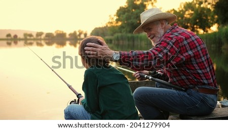Back view of the senior man chatting with his grandson and stocking him at the head while sitting with the fishing roads and fishing at the pier. Family concept