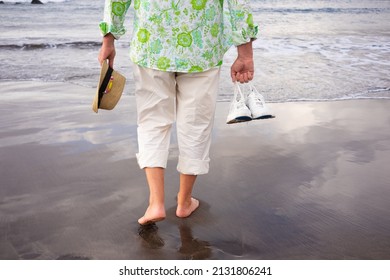 Back View Of Senior Man Walking On The Beach Barefoot Holding Shoes And Hat In Hands Enjoying Vacation Walking On The Sand. Concept Of Relaxed And Free Retiree