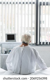 Back View Of Senior Patient In Gown Sitting On Bed In Hospital Ward