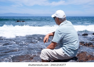 Back view of senior relaxed man barefoot wearing sunglasses and cap sitting on a rocky beach admiring the sea waves. Travel vacation leisure activity concept - Powered by Shutterstock