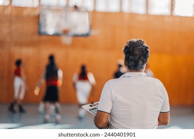 Back view of a senior trainer standing on court indoors and watching basketball training.Rear view of a professional senior basketball coach watching basketball players on training.Basketball training - Powered by Shutterstock