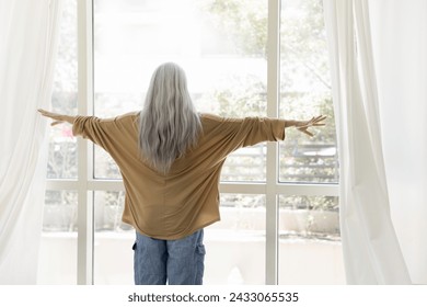 Back view of senior woman with long naturally grey hair in casual opening drapes, parting white transparent curtains, veils at large window, enjoying view of terrace view - Powered by Shutterstock